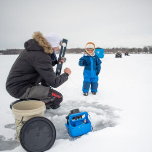 Parent and child in snow gear on frozen lake to ice fish.