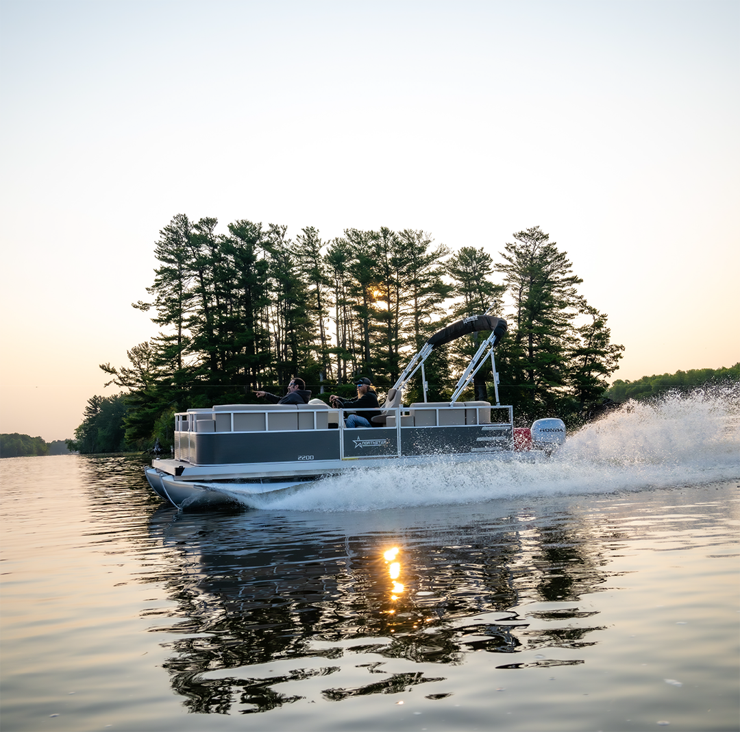 A Cruise Water Pontoon drives past a pine tree covered island at sunset.